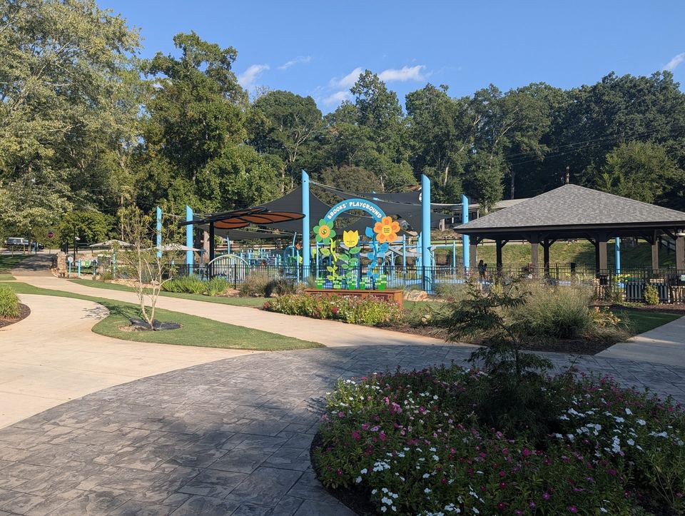 A colorful playground and picnic shelters beyond a paved plaza and flower bed.