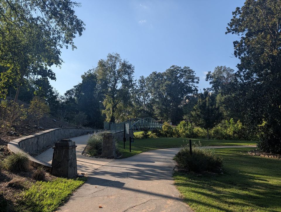 A curving paved path between stone pillars leading toward a footbridge in the trees.