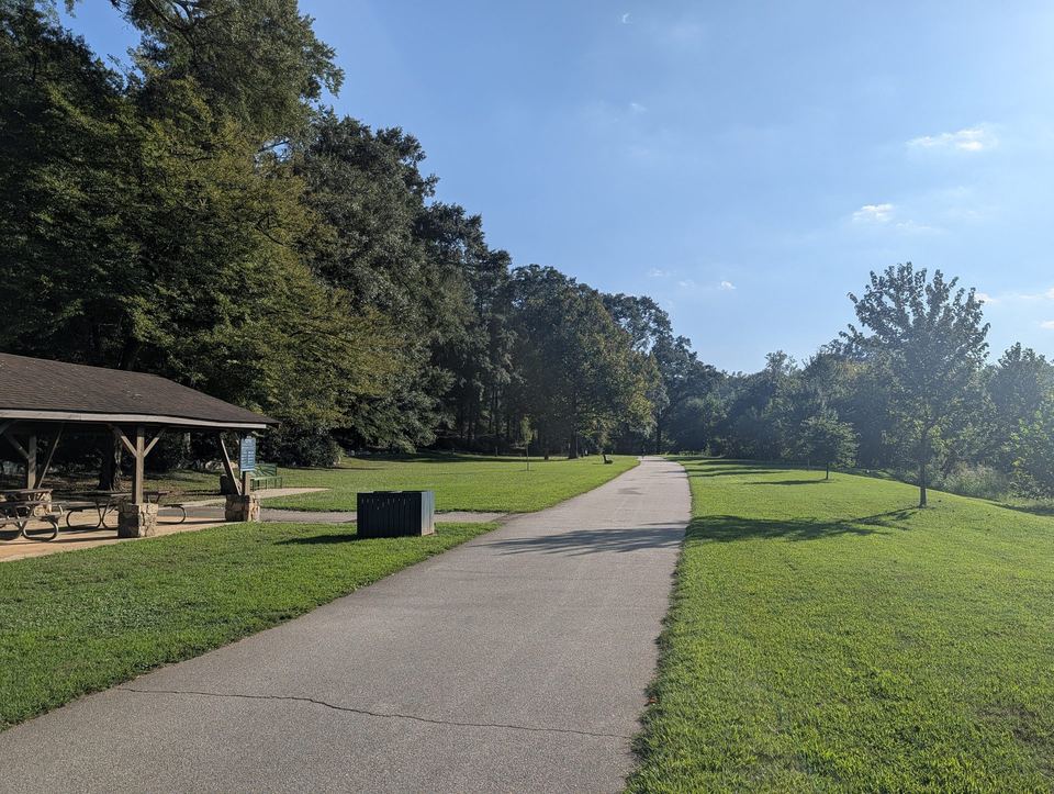 A paved path running between lawns past a picnic shelter and tall trees.